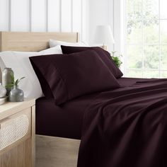 a bed with purple sheets and pillows in front of a large window, next to a night stand