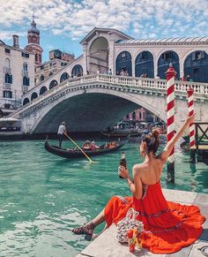 a woman in an orange dress sitting on the edge of a bridge with gondola passing by