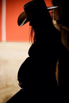 the silhouette of a pregnant woman wearing a cowboy hat and sitting against a red wall