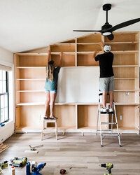 two people standing on ladders in an unfinished room