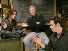 three men sitting in front of a blackboard with writing on it and one man holding his hands out