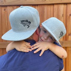 a father hugging his son in front of a wooden fence wearing a hat with fish embroidered on it