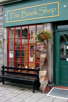 the book shop has many books stacked up on it's front window and there is a wooden bench outside