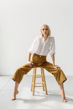 a woman sitting on top of a wooden stool in front of a white wall with her legs crossed