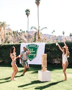 two women and a man playing with a giant block tower in front of palm trees