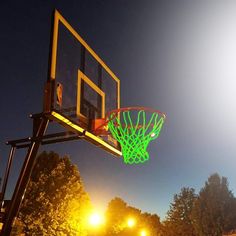 a basketball hoop is lit up at night with the sun shining on trees in the background