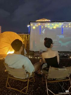 two people sitting in lawn chairs watching a movie on the screen at night with umbrellas