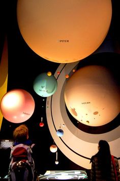 people are looking at the planets on display in a museum with lights hanging from it's ceiling