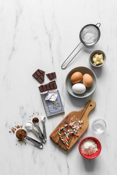 ingredients to make christmas cookies laid out on a marble counter top, including eggs, chocolate and candy canes