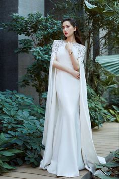 a woman in a white gown and cape standing on a wooden deck surrounded by greenery