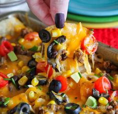 a hand holding a piece of nacho with black olives, corn and tomatoes