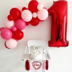 a chair and balloon garland on the wall for a one year birthday party with red, white and pink balloons