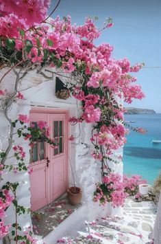 a pink door is surrounded by flowers on the side of a white building with blue water in the background