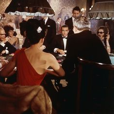 a woman in a red dress sitting at a table surrounded by other people and wearing tuxedos