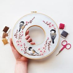 a hand is holding up a embroidery hoop with birds on it and thread spools next to the hoop