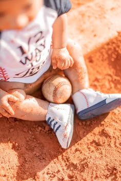 a young boy sitting on top of a baseball field holding a ball and wearing blue shoes