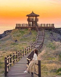 a woman sitting on top of a wooden staircase next to the ocean with a gazebo in the background