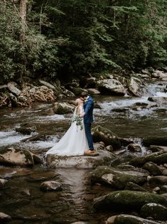 a bride and groom are standing on rocks in the middle of a stream, surrounded by trees