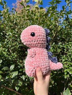 a hand holding a pink and blue crocheted stuffed animal in front of some bushes