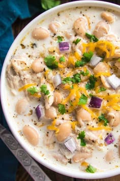 a white bowl filled with chicken and bean soup on top of a wooden table next to a spoon