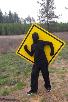 a man holding a large yellow sign in the middle of a grass covered field with trees behind him