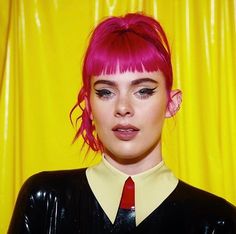 a woman with bright pink hair wearing a black shirt and red tie, standing in front of a yellow curtain