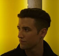 a young man in a black jacket looks off into the distance while standing against a yellow wall