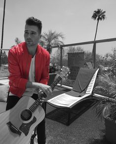 a man holding an acoustic guitar in front of a bench with palm trees and chairs behind him