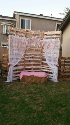 a bed made out of wood pallets in the grass next to a building with a pink curtain over it