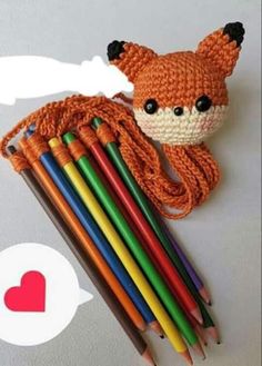 an orange crocheted fox head next to several colored pencils and a red heart