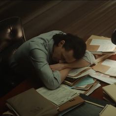 a man sitting at a desk covered in papers and paperwork with his head on his hands