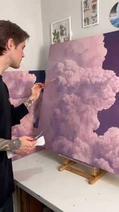 a man in black shirt painting clouds with purple and white paint on canvases,