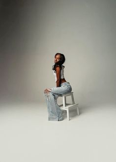 a woman sitting on top of a white chair in front of a gray background with her legs crossed