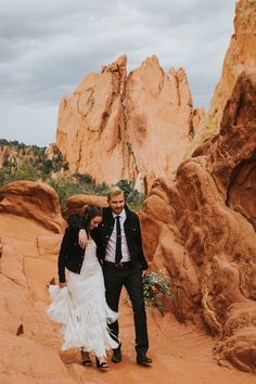 a bride and groom are walking through the desert with rocks in the backgroud