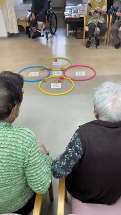 two elderly women sitting in chairs looking at an object on the floor with people watching