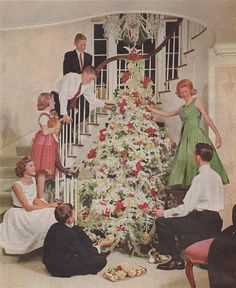 a group of people sitting around a christmas tree in front of a staircase with the man and woman decorating it