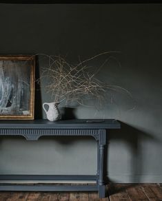 a painting and vase on a table in front of a gray wall with wood flooring