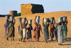 a group of people carrying buckets on their heads in the desert with sand dunes behind them