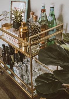 a gold bar cart filled with bottles and glasses on top of a wooden floor next to a potted plant