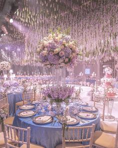 a table is set with blue linens and purple flowers hanging from the ceiling above it