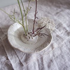 two flowers are in a white bowl on a linen tablecloth, one is sprouting from the top