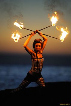 a woman holding two torches over her head with fire in the air above her head