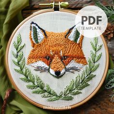 an embroidered fox head on a wooden surface with pine needles and green leaves around it