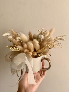 a hand holding a coffee mug with dried flowers in it and a ribbon around the handle