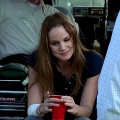 a woman sitting on the ground holding a red cup and looking at her cell phone