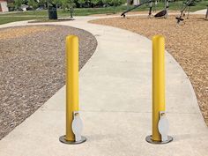 two yellow poles on the side of a sidewalk near a park with children's swings