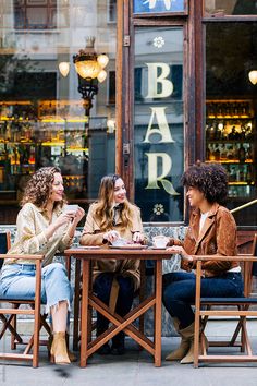 three women sitting at a table in front of a bar talking to eachother