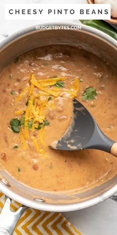 a pot filled with cheese and beans on top of a yellow table cloth next to a wooden spoon