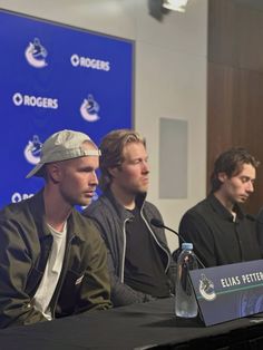 three men sitting at a table with microphones in front of them and a sign behind them