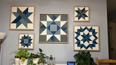 four framed quilts hang on the wall above a table with a laptop and potted plants